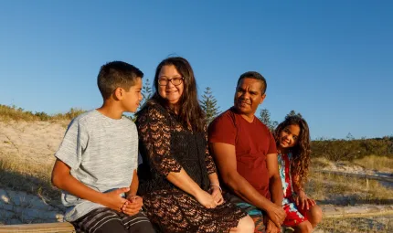 In the image is a family of four. A mum and dad with son and daughter sitting on a bench talking to each other outdoors.