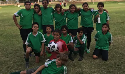 On a grass field is a group of 14 Aboriginal youth in green soccer jerseys and black shorts. The back row is standing, the middle row is kneeling and one young man lays down in front of the rest. One young man is in a red shirt and holds a soccer ball. In