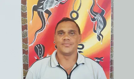 Young Aboriginal man in pale blue shirt stands in front of Indigenous artwork displaying dark brown and white animals on a red and yellow background.