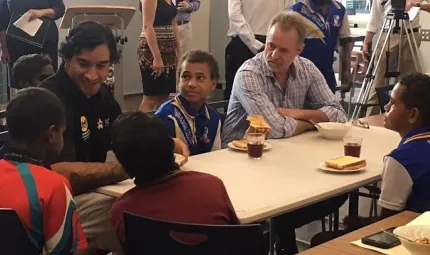 An Indigenous man and non-Indigenous man sit at a table with Indigenous children while other adults and an Indigenous boy stand in the background.