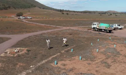 Two men in beige workwear stand on open ground near some newly planted saplings held up with posts and feral animal protection. In the background are trucks, a hill and blue sky.