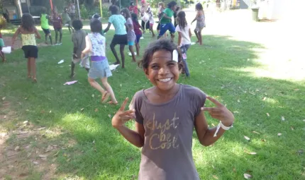 Young Aboriginal girl smiling at camera and other children in background. All are playing on the grass.