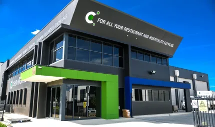 Image is a modern black building with windows and a green entrance. At right are large roller doors. The entire foreground is a concrete slab or driveway.