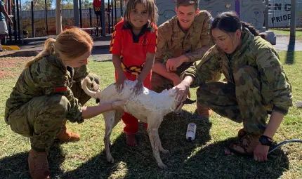 Young Aboriginal girl in red school uniform and three soldiers (2 women and 1 man) surround a white dog on a grassed area. One soldier holds a hose. In the background is a playground.