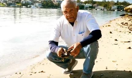 Elderly Aboriginal man in white shirt and jeans squats next to a river on a sandy shore. In the background on the other side of the river are houses and trees.