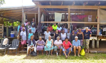 Group of Aboriginal women and children dressed in colourful clothing stand in front of a grey and blue building on a path with grass in the foreground.