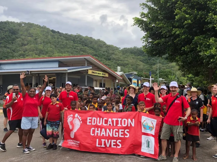 A group of men, women and children in red school polo shirt with a crowd of Indigenous children behind red banner with white writing on banner ‘Education Changes Lives.’