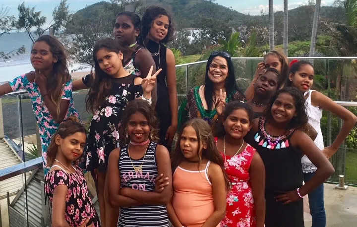 Aboriginal woman with a group of 12 year 5 and 6 Aboriginal girls. They are wearing colourful dresses, and a standing on a balcony with a beach behind them.