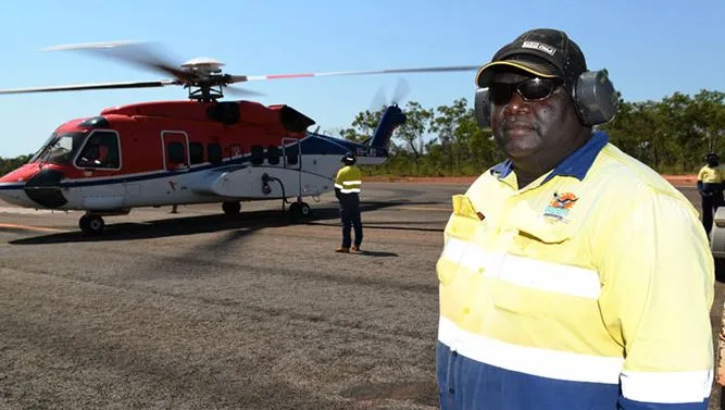 A man in work wear and including safety headphones and a cap stands on an aircraft tarmac. In the background are other men and a large helicopter. Behind that is bush land.