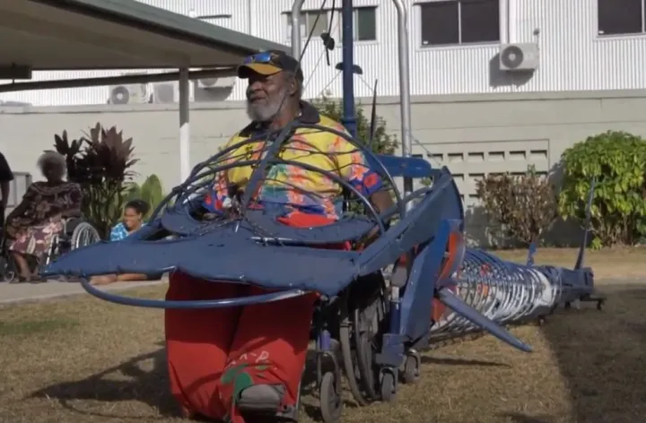 An elderly man sits in a wheel chair that forms part of a long metal contraption in the shape of a hammerhead shark. In the background are two women, grass, shrubbery and a building.