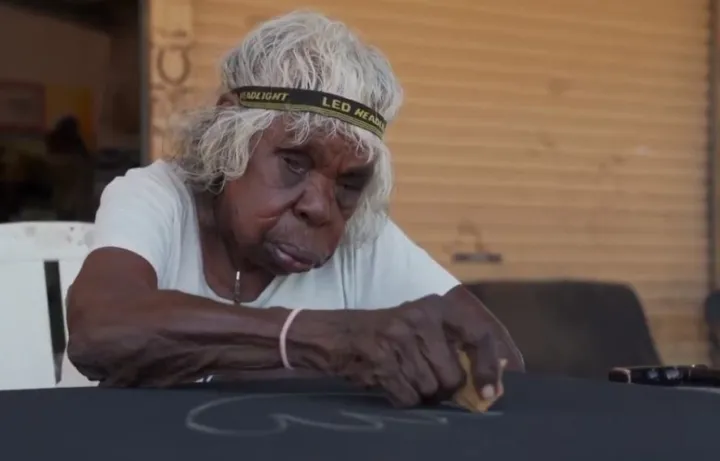 An elderly Aboriginal woman wearing a headband around her grey hair sits at a table and sketches on a black canvas.