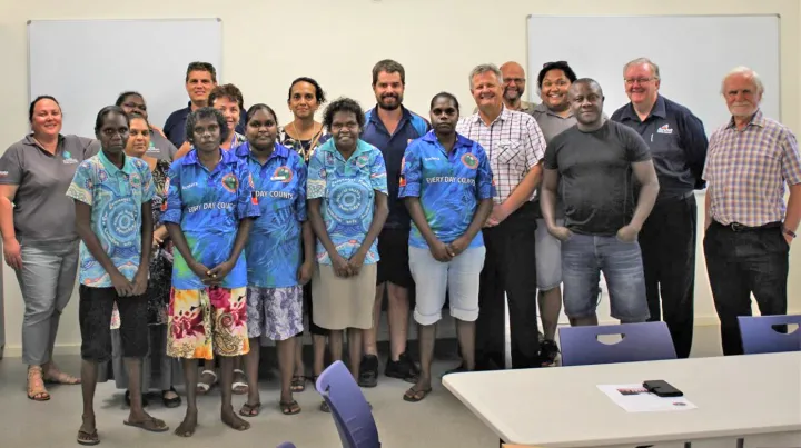 Large group of Indigenous and non-Indigenous men and women dressed in casual clothing in a room with beige carpet. In the background is a pale yellow wall and two whiteboards. In the foreground is a white table and blue chairs.