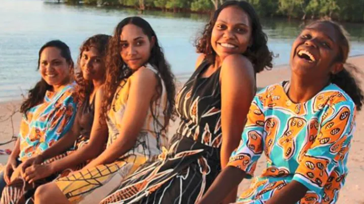 Five Indigenous women wearing colourful clothing sit in a line on a beach with water and trees in the background.