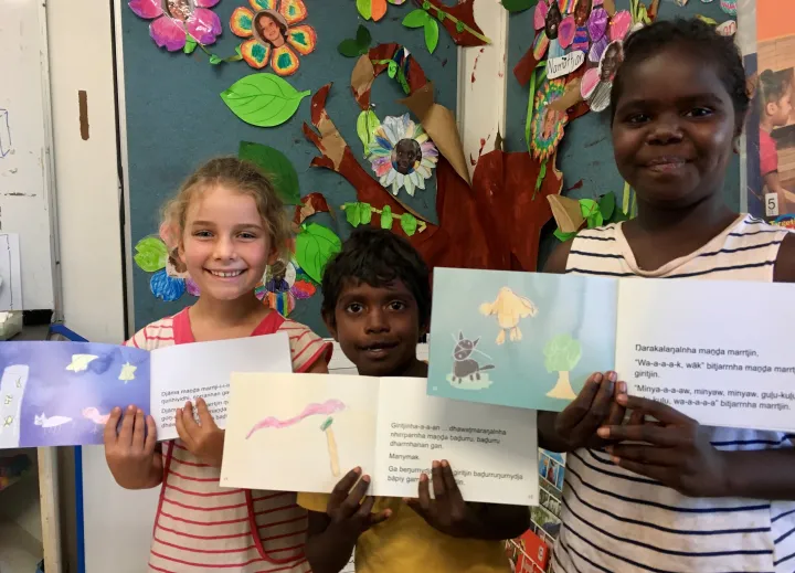 Three children stand in front of a colourful artistic display. Each holds a children’s book open to a two page spread with paintings on the left and text on the right.