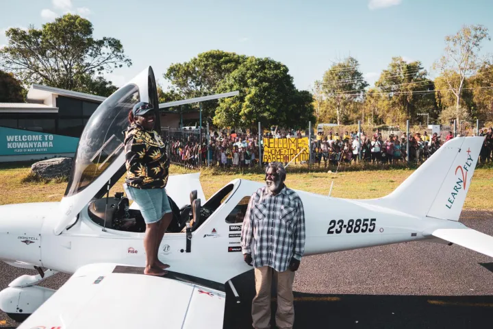 A young Indigenous woman, Tyeisha Clarke, is standing in bare feet on the wing of a light aeroplane, after exiting the cockpit. She is pictured with her father John. The Kowanyama community are standing behind a secure fence away from the airstrip.