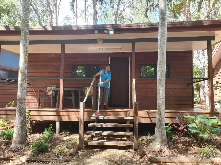 A woman in work wear sweeps the deck in front of a wooden cabin. At the front are two palm trees, some foliage and steps leading up to the cabin entrance. In the background are more trees.