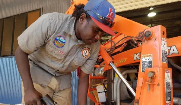 Young Aboriginal man dressed in work wear holds a tool to pump up a black tyre on a heavy machine.