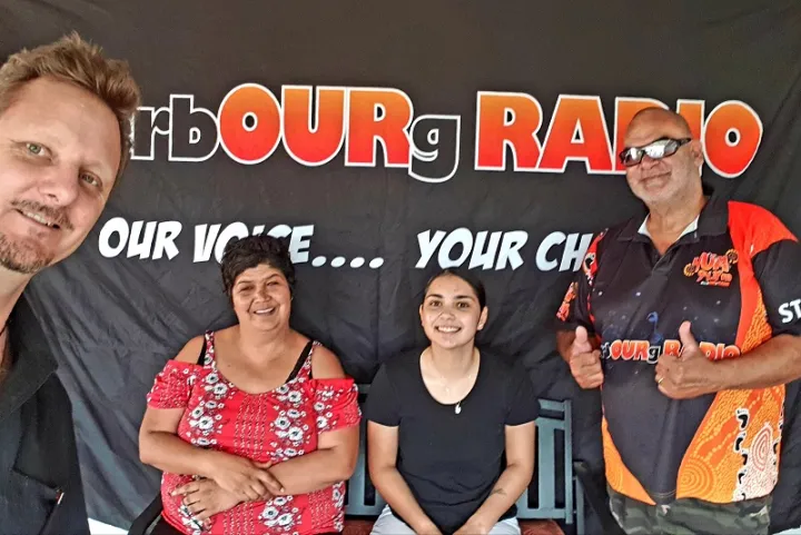 Two women sit in front of a black background with two men standing either side of them. Words on the background are Cherbourg Radio Our Voice…Your CH