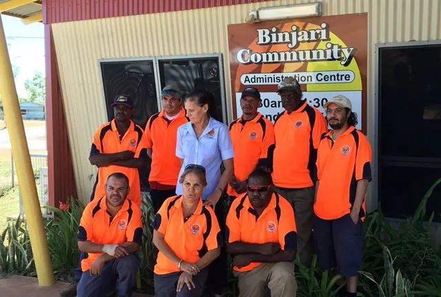 Two rows of nine Indigenous persons dressed in orange shirts (one in blue) stand and kneel in front of a yellow building with a sign on the wall which says, Binjari Community Administration Centre.