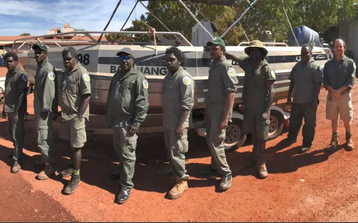 Group of men and a woman dressed in ranger uniforms stand in front of a boat. In the background are trees.