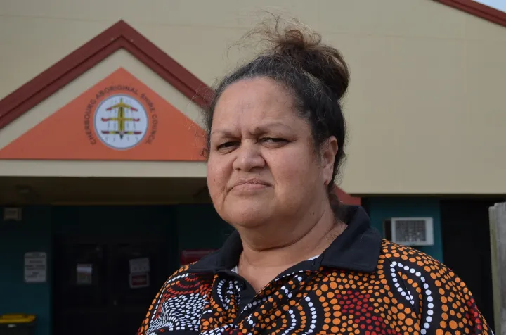 An Aboriginal woman with her hair up and wearing a polo shirt with Indigenous design stands in front of a building.