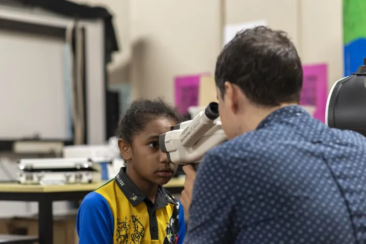 A young child looks into a piece of eye testing apparatus while a man looks into the other end. In the background is a table and other equipment.