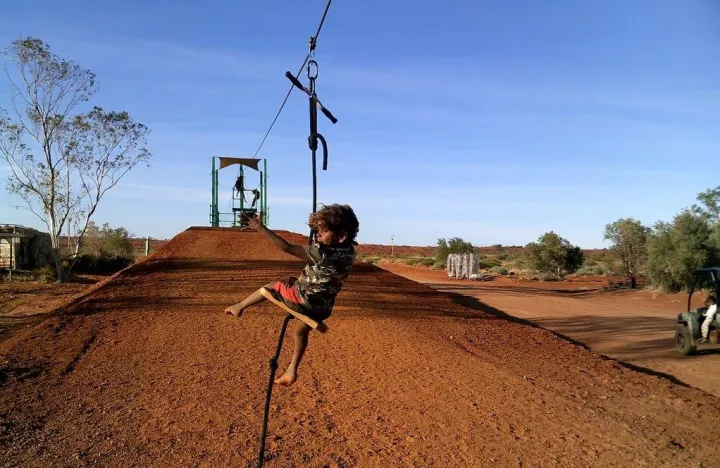 Aboriginal child on a cable suspended over brown soil. In the background is bare soil and some trees.