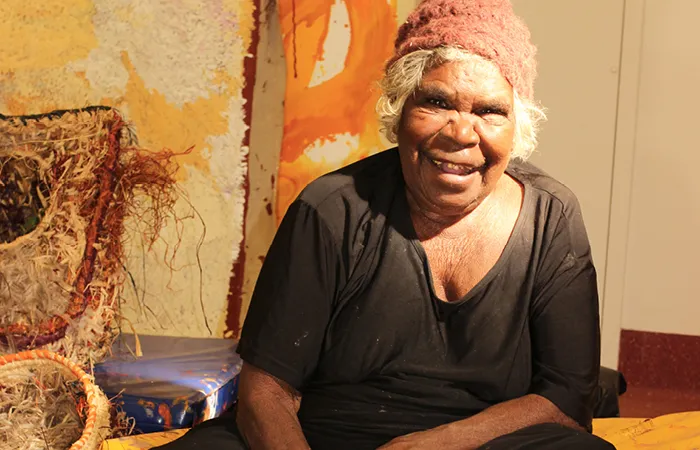 An elderly woman in black dress and reddish beanie is sitting down. In front of her is artwork featuring some birds. In the background is more art and a beige coloured wall.