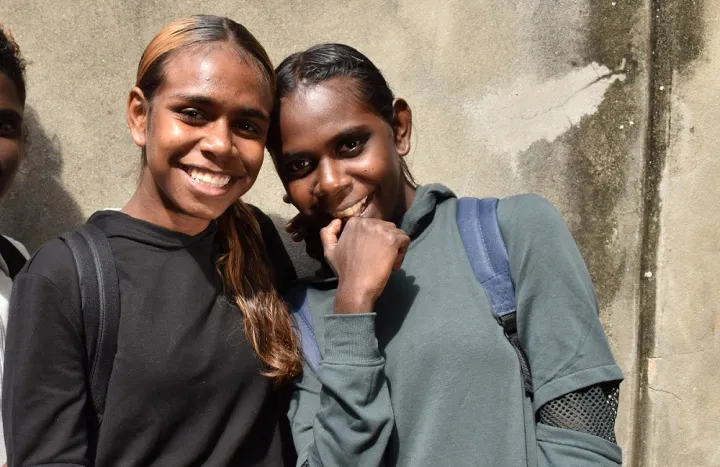 Two Aboriginal young women standing side by side and heads together. The one on the left is wearing a black top and the other an olive coloured top. In the background is a concrete wall.