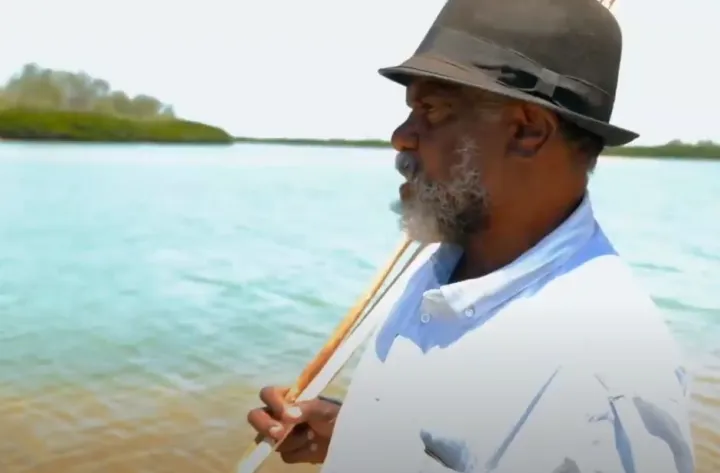 An Indigenous man in hat and pale shirt holds a spear over his shoulder. In the background is water and foliage.