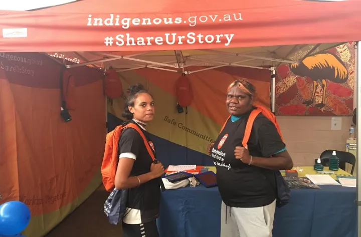 Two young Indigenous women wearing orange backpacks and dressed in Batchelor Institute shirts stand in front of a blue table topped with papers and pamphlets set up in a stall displaying the words: indigenous.gov.au #ShareUrStory.