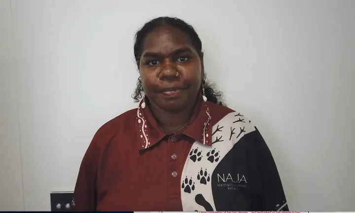 An Aboriginal woman with hair tied back wears an ochre red, white and black shirt with Aboriginal designs over the left shoulder. In the background is a white wall.