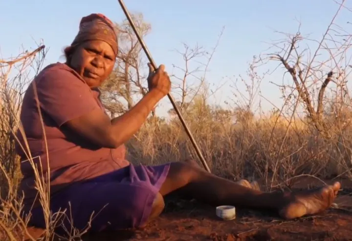 A mature Aboriginal woman dressed in top and skirt sits on red soil in amongst long grass. She holds a metal rod in her right hand.