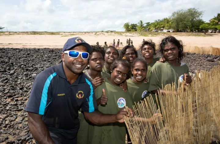 Aboriginal man in blue shirt and cap with Aboriginal youth in green shirts stand on rocks at a beach with sand and trees in the background. He supports an upright bamboo barrier.