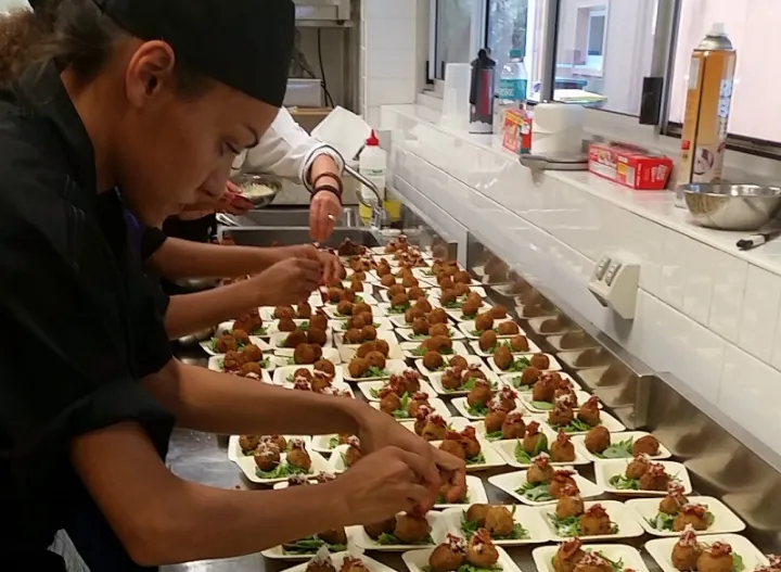 Indigenous woman dressed in black prepares multiple food dishes on stainless steel bench with fellow chefs in the background.