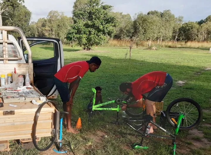 A man in red shirt bends over a green pushbike laying on grass. Watching at left is another man in red shirt and behind is a young boy sitting and watching. At left is a truck and in the background is grass and trees.