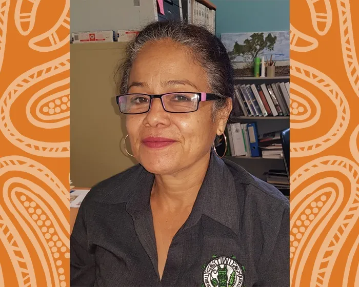 Indigenous woman with hair drawn back and wearing glasses and a dark shirt. Behind her is shelving and other office content.