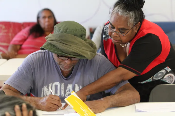 Indigenous man with green hat and grey t-shirt writes on a paper as an Indigenous woman in red and black apparel points at the paper. Another Indigenous woman sits on a couch in the background.