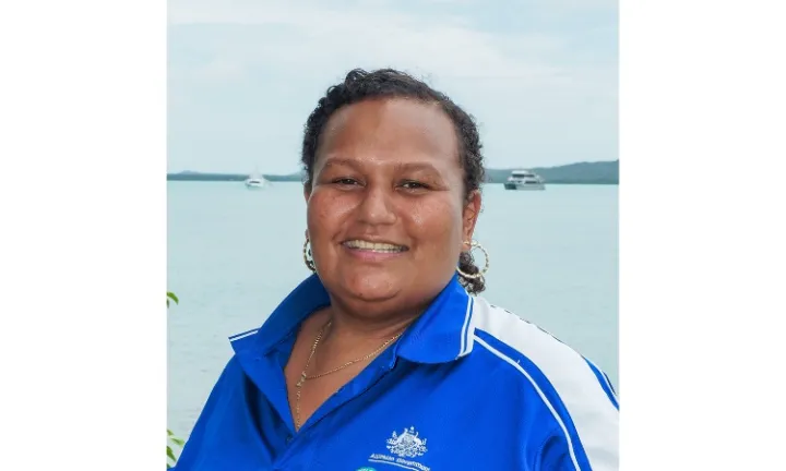 A woman in blue and white polo shirt faces the camera. In the background is blue water, two boats and some land.