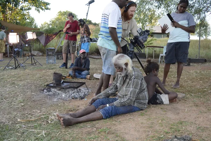 Elderly Aboriginal man sits back to back with young Aboriginal boy on grassy ground. Immediately behind them are three film-makers and further back are more Aboriginal and non-Aboriginal film makers another Aboriginal actor, lights, trees and grass.