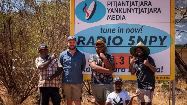 Five men in casual wear stand or sit in front of a large multi-coloured sign with a logo at top left and the following text: Pitjantjatjara Yankunytjatjara Media. Tune in now Radio 5NPY. In the background are trees, grass and a building.