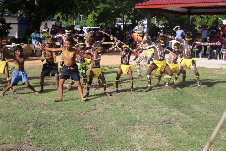 A group of Indigenous children with white ceremonial paint on their bodies wearing shorts or yellow coverings dance on green grass watched by a crowd of onlookers.