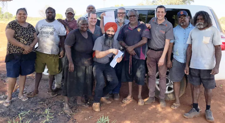 There are 12 adults standing together and facing the camera. They are outside on a sunny day, standing in the shade, in front of a white van.