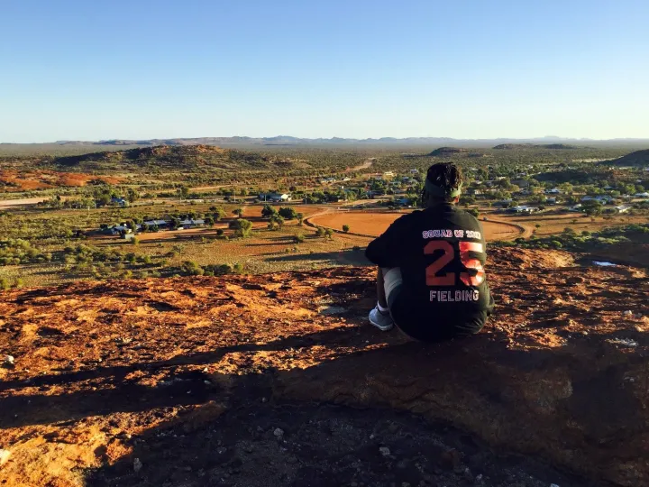 Aboriginal woman sits on a hill overlooking a small town surrounded by low hills in an arid landscape. On the back of her dark top is ‘Squad of 2017, 25, Fielding’.