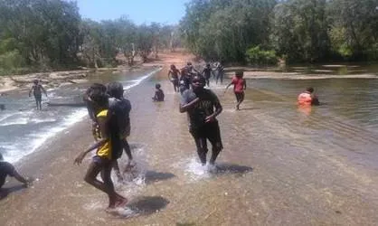 Several youth stand on a roadway over which flows a river. At left are rocks in the stream and in the background is a dirt road, trees and blue sky.