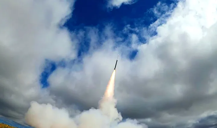 A rocket with a gas trail behind it heads toward a sky with a bit of blue but mainly cloudy.