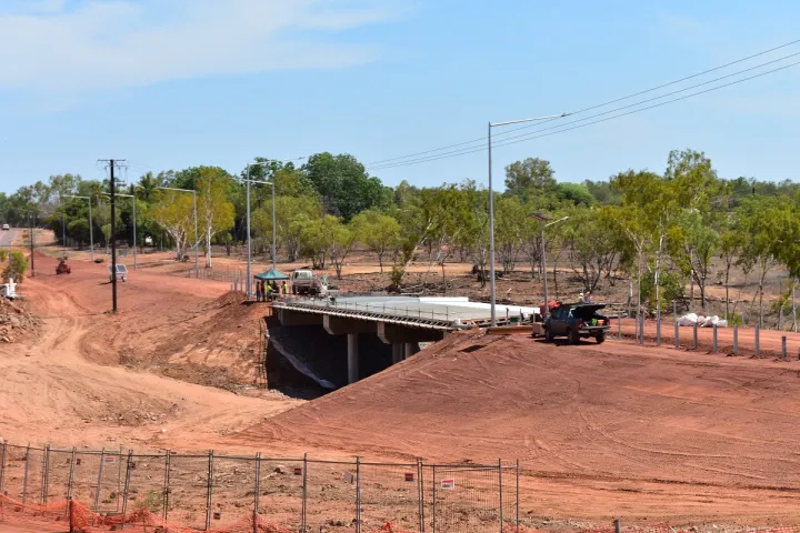 In the foreground is ochre coloured soil behind which is a concrete bridge with vehicles nearby and in the background are trees and blue sky.