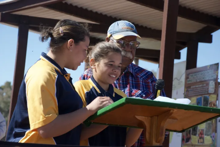 Two young Aboriginal girls dressed in blue and yellow polo shirts stand at a podium with an elderly Aboriginal man in blue cap and blue and red shirt. In the background is a small metal shelter.