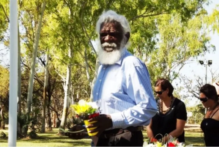 Elderly Aboriginal man with white hair and beard and dressed in pale blue shirt is holding a pot of flowers. In the background are two women dressed in black and a line of tall leafy trees.