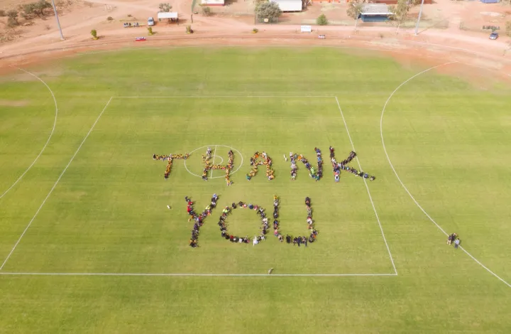 A bird’s eye view of people on a green oval. They stand close together and form the words ‘Thank you’. In the background is a brown road, buildings and more people.